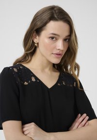 Blouse noire à manches courtes, avec un tissu texturé et des détails en dentelle aux épaules. Design en V. Boucles d'oreilles dorées en accent.