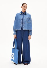 Denim jacket with buttons, wide-leg denim pants, and a blue tote bag featuring a white graphic. Model wears black sandals.