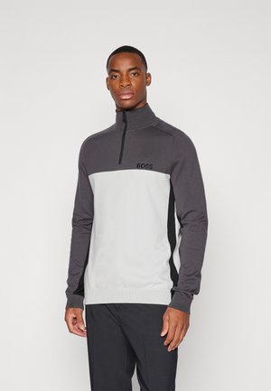 Suéter con cremallera en tonos gris y blanco, con un cuello alto y un logo en el pecho, además de detalles laterales en negro. Fabricado con un tejido de punto suave.