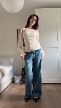 Jeune femme posant à l'intérieur, portant un haut beige transparent à manches longues, un jean bleu à jambes larges et des chaussures noires à bouts pointus, dans une chambre.