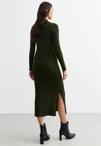 Robe en tricot vert foncé avec texture côtelée, manches longues et fente latérale ; associée à des bottines noires.