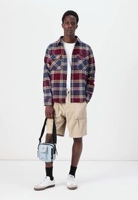 Flannelskjorte i marineblå, bordeaux og creme-rutet; hvid t-shirt; beige shorts; blå crossbody taske; hvide sneakers med brune detaljer.