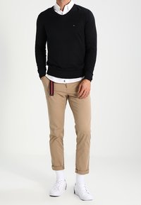 Schwarzer V-Ausschnitt-Pullover über einem weißen Hemd, khakifarbene Chinos und weiße Sneakers. Mit hochgekrempelten Hosenbeinen und kleinem Logodetail auf dem Pullover.