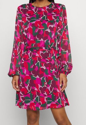 Kvinde iført langærmet, knælængde kjole med et lyst rødt, pink, grønt og lilla abstrakt blomstermønster og bælte med bindebånd i taljen.