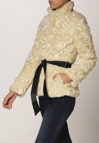 Syntetisk pels cremefarvet jakke med høj krave, lange ærmer og en sort taljebind. Har en blød tekstur og to forlommer.