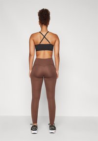 Soutien-gorge de sport noir avec des bretelles croisées, leggings taille haute marron, et chaussures de sport noires. Texture lisse et design ajusté.