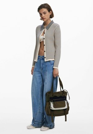 Striped cardigan in cream and navy, paired with loose denim jeans. A multi-pocketed olive bag with black and cream accents is held by the model.