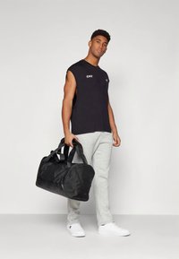 Bolsa deportiva negra con doble asa y base curva, llevada por una persona con camiseta negra sin mangas y pantalones deportivos grises, de pie frente a un fondo liso.