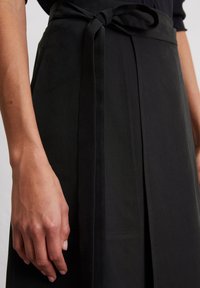 Gonna nera con vita legata, pieghe e una texture liscia. Il tessuto appare leggero con una lucentezza sottile e linee pulite.
