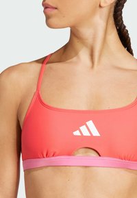 Reggiseno sportivo di un rosa brillante con un design a taglio, caratterizzato da un cinturino rosa a contrasto e un logo bianco. Texture liscia, spalline regolabili.