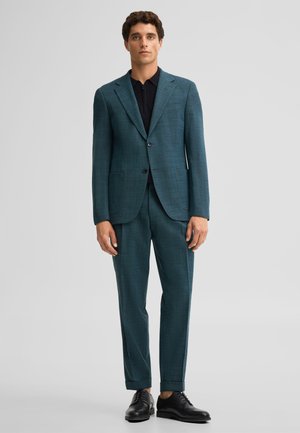 Uomo che indossa un abito verde petrolio scuro con scarpe eleganti nere, in piedi davanti a uno sfondo grigio chiaro uniforme.