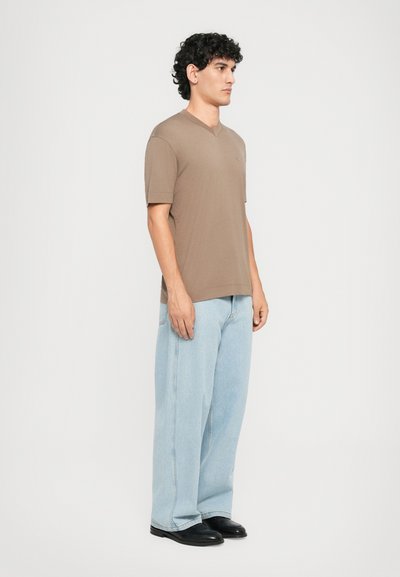 Homme se tenant debout dans un t-shirt V-neck beige, un jean large bleu clair et des chaussures noires, contre un fond blanc uni, tourné vers la droite.