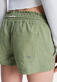 Shorts en lin verts avec une ceinture élastique, dotés de deux poches arrière et d'un petit logo sur la poche droite.