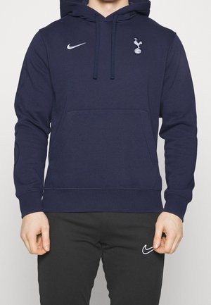 Mand iført navyblå Nike-hættetrøje med Tottenham Hotspur-logo og sorte Nike-bukser, stående mod en ensfarvet baggrund.