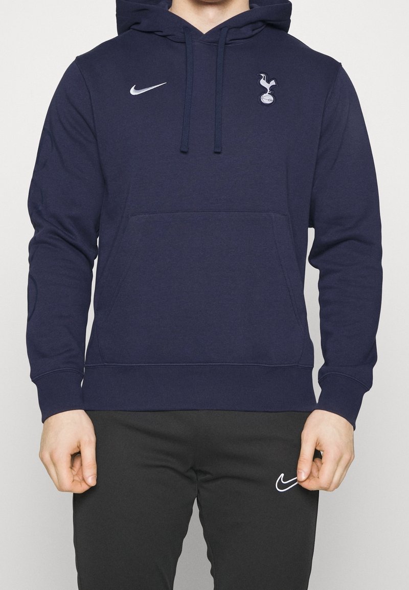 Homme portant un sweat à capuche Nike bleu marine avec le logo de Tottenham Hotspur et un pantalon Nike noir, debout devant un fond uni.