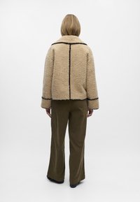 Beige Fake-Shearling-Jacke mit schwarzem Besatz, ausgestattet mit einem Kragen und lockerer Passform. Kombiniert mit olivgrünen, weit geschnittenen Hosen.