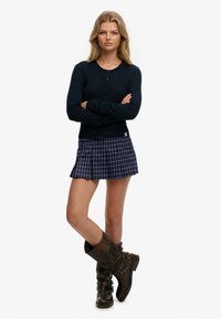 Marineblå ribstrikket cardigan med knaplukning, kombineret med en plisseret ternet mininederdel i blå og hvid, suppleret med brune knælange støvler.