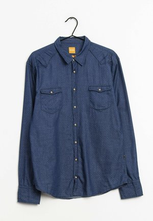 Langärmliges blaues Denim-Hemd mit Knöpfen und zwei geknöpften Brusttaschen, hängend an einem schwarzen Kleiderbügel vor weißem Hintergrund.