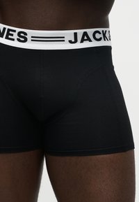 Boxers noirs ajustés sur une personne avec une ceinture blanche affichant un texte noir partiel "JACK & JONES" sur un fond uni.