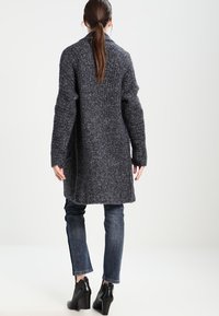 Długi, ciemnoszary płaszcz z mieszanki wełny o teksturowanym wykończeniu i szerokich rękawach. Noszony na niebieskie dżinsy i czarne botki na obcasie.