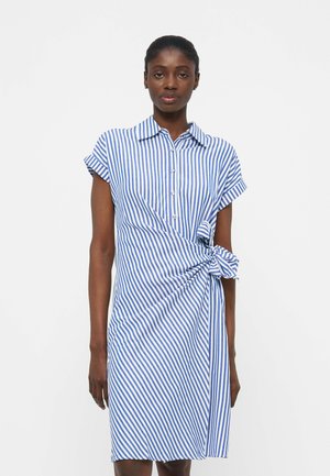 Čierna žena v modrom a bielom krátkom rukávovom šate s vertikálnymi pruhmi, golierom a detailom viazaného uzla na pravej strane.