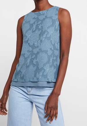 Sleeveless blue top with raised floral embroidery worn by person with medium-dark skin and light blue jeans against white background.