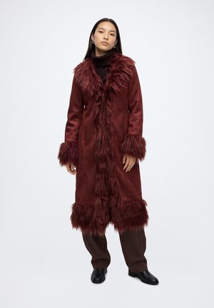 Femme portant un long manteau bordeaux avec une bordure en fausse fourrure au col, aux poignets et à l'ourlet, associé à un pantalon foncé et des chaussures noires, debout sur fond blanc.