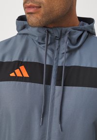 Grå jakke med hætte, sort og orange vandret stribe, lynlåslukning og snoretræk til justering. Glat tekstur og sporty design.