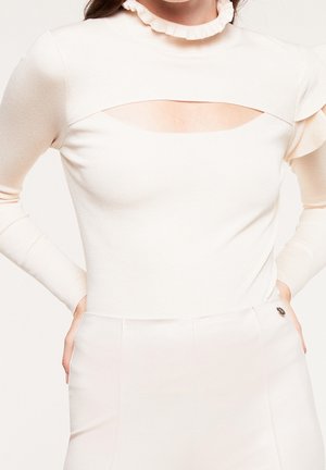 Femme portant un haut crème à manches longues avec un col à volants et une découpe horizontale au-dessus de la poitrine, rentrée dans un pantalon taille haute.