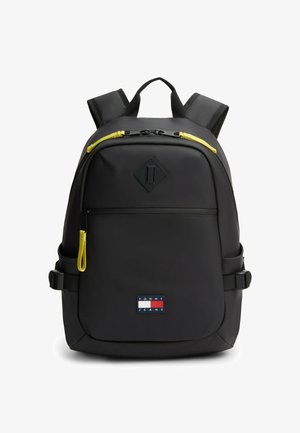 Czarny plecak z żółtymi suwakami, przednią kieszenią na zamek, wyściełanymi paskami na ramiona oraz naszywką z logo Tommy Jeans na dolnej przedniej części.