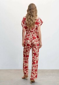Mujer con cabello largo y ondulado lleva un mono beige con estampado floral rojo y sandalias de tacón beige, de pie frente a un fondo liso.