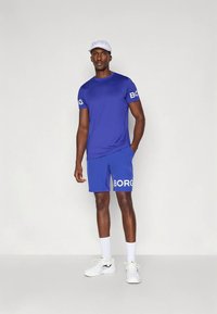 Άνδρας που φοράει μπλε αθλητικό T-shirt και σορτς με το κείμενο "BORG", λευκό καπέλο, λευκές κάλτσες και λευκά αθλητικά παπούτσια, στέκεται μπροστά σε απλό φόντο.