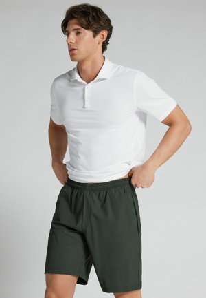 Mann passt dunkelgrüne Shorts an, während er ein weißes kurzärmeliges Polohemd vor einfarbigem Hintergrund trägt.