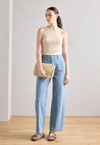 Mujer con top sin mangas beige, pantalones vaqueros de pierna ancha azul claro, sandalias marrones con hebilla, sosteniendo un clutch de fibra tejida, de pie sobre un suelo de madera contra una pared blanca.