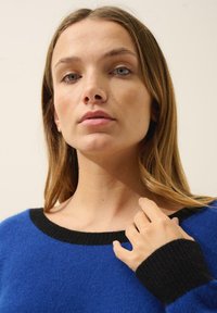 Modrý svetr s černým žebrovaným výstřihem a manžetami rukávů. Měkká textura, uvolněný střih, široký výstřih a dlouhé rukávy.