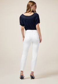 Jeans bianchi attillati con vita alta, abbinati a una blusa navy con scollatura bordata in pizzo e maniche a sbuffo. Sandali con tacco completano l'outfit.