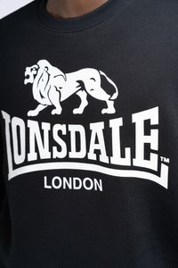 Fekete pulóver kerek nyakkivágással, nagy fehér oroszlán grafikával és vastag, kontrasztos fehér "LONSDALE LONDON" felirattal.