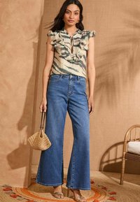 Blusa floreale a volant in crema, verde e rosa, abbinata a jeans in denim blu a gamba larga. Gli accessori includono una borsa intrecciata e sandali metallici.