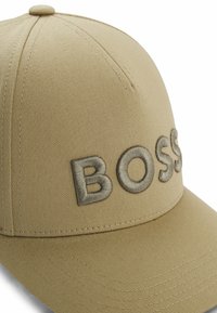 Khaki baseball cap featuring raised, silver embroidered "BOSS" text on the front. Smooth fabric with reinforced stitching and a rounded crown.