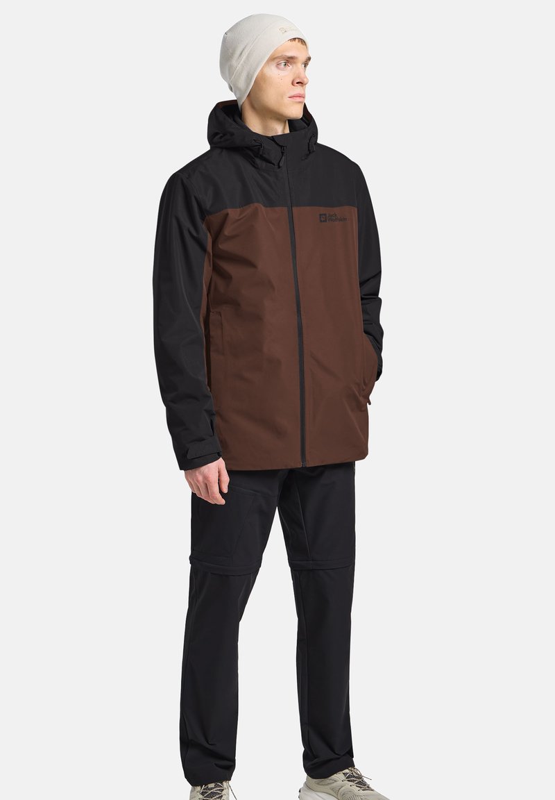 Giacca impermeabile marrone e nera con cappuccio, chiusura con zip e tasche laterali, abbinata a pantaloni neri e scarpe chiare.