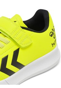 Neon gul sneaker med svarta accenter, som har en texturerad yta, kardborreband och en vit gummisula med kantiga designelement.