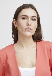 Koraalkleurige blazer met een zachte textuur, open voorkant en brede revers, gedragen over een witte tanktop. Gouden creolen voegen een metallic accent toe.
