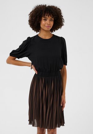 Donna con capelli ricci che indossa una maglia nera con maniche a sbuffo e una gonna plissettata trasparente con strisce verticali marroni e nere, in piedi con una mano sui fianchi.