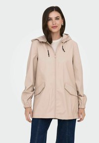 Chaqueta impermeable de un color beige claro con capucha, cremallera frontal, puños elásticos y dos bolsillos laterales. Textura suave, diseño informal, de longitud media.