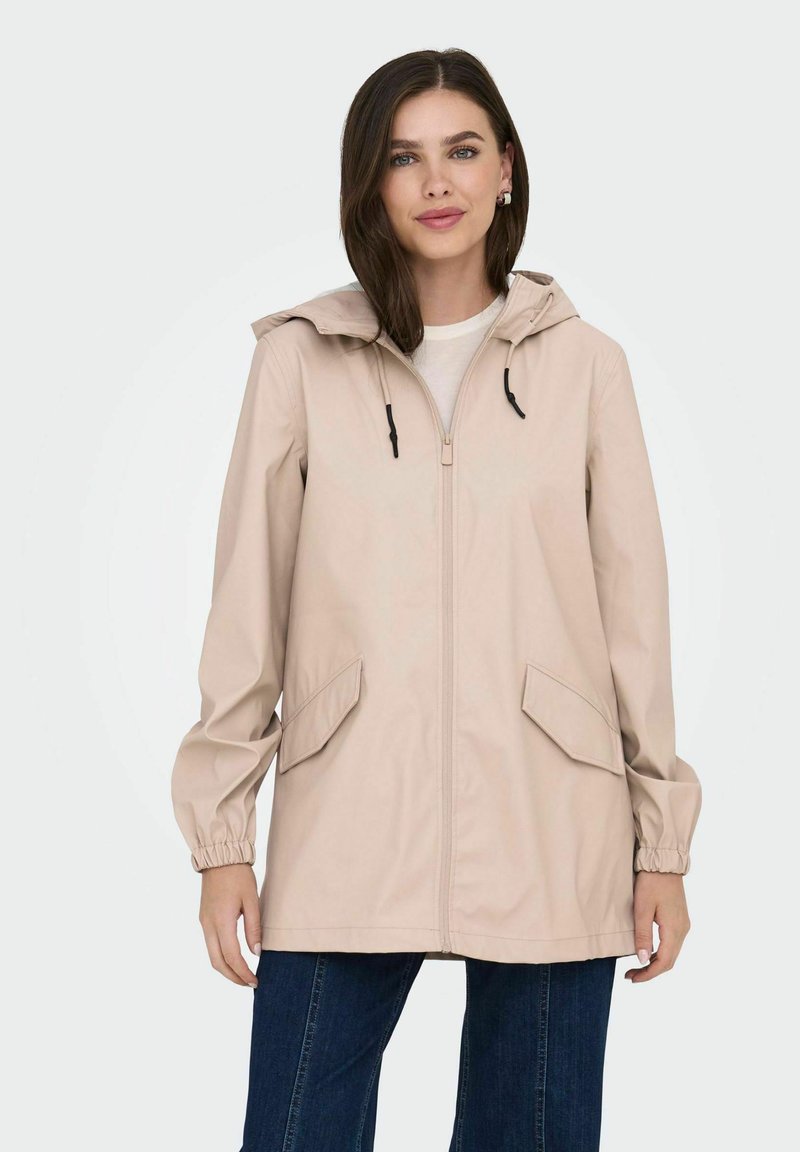 Chaqueta impermeable de un color beige claro con capucha, cremallera frontal, puños elásticos y dos bolsillos laterales. Textura suave, diseño informal, de longitud media.
