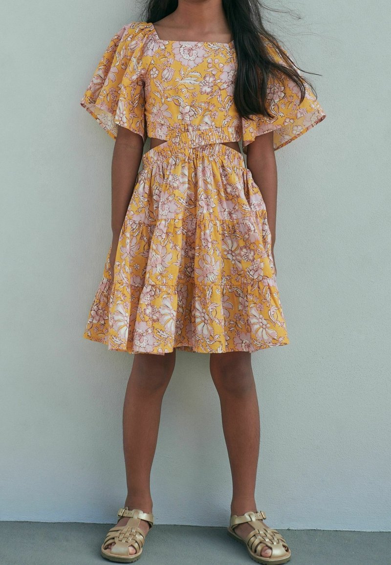 Fille portant une robe jaune à imprimé floral avec des manches courtes bouffantes et des sandales dorées, debout devant un mur clair uni.