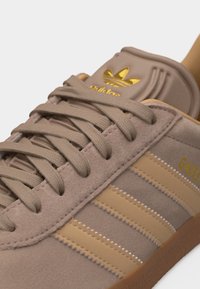 Close-up van een beige Adidas Gazelle sneaker met suède materiaal, beige veters, drie beige strepen en een gouden Adidas-logo op de tong.