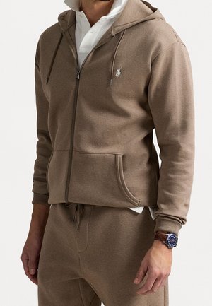 Mężczyzna ubrany w brązową bluzę z kapturem na suwak oraz dopasowane spodnie dresowe, pod spodem biała koszulka polo oraz zegarek na rękę z niebieską tarczą i brązowym skórzanym paskiem.