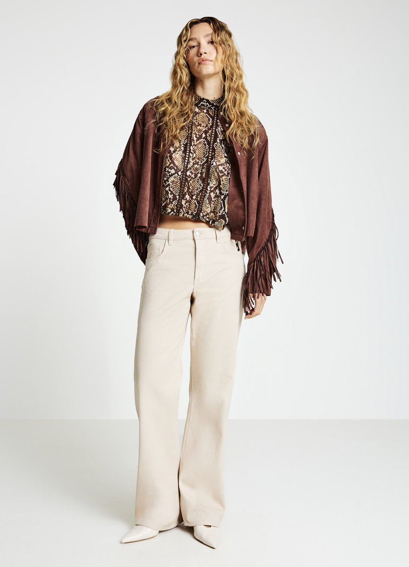 Veste courte en suède marron avec franges, haut à imprimé serpent, pantalon beige à jambes larges, et escarpins blancs à bout pointu, le tout sur un fond clair.