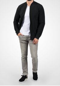 Schwarze Bomberjacke über einem weißen T-Shirt, kombiniert mit hellgrauen Jeans und schwarzen Turnschuhen. Verfügt über einen Reißverschluss, gerippte Bündchen und ein sportliches Design.